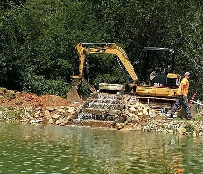 DeBord & Son Construction | working on cleaning out shoreline of pond with a bobcat in a wooded area